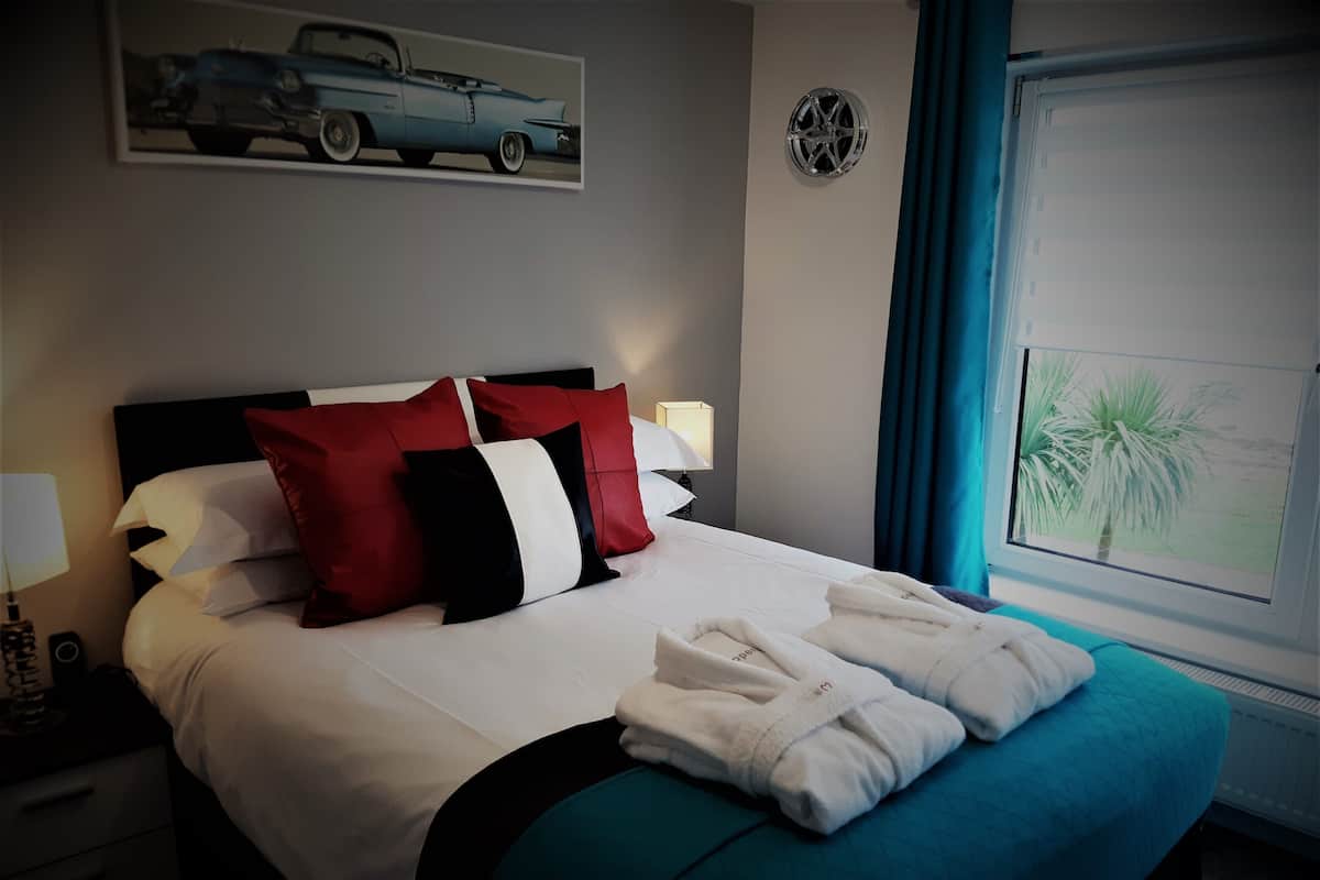 A neatly made bed with white and red pillows, a teal bedspread, and two white towels.