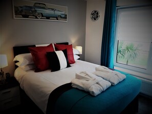 A neatly made bed with white and red pillows, a teal bedspread, and two white towels. - Cadillac Kustomz Guest House (Rothesay)
