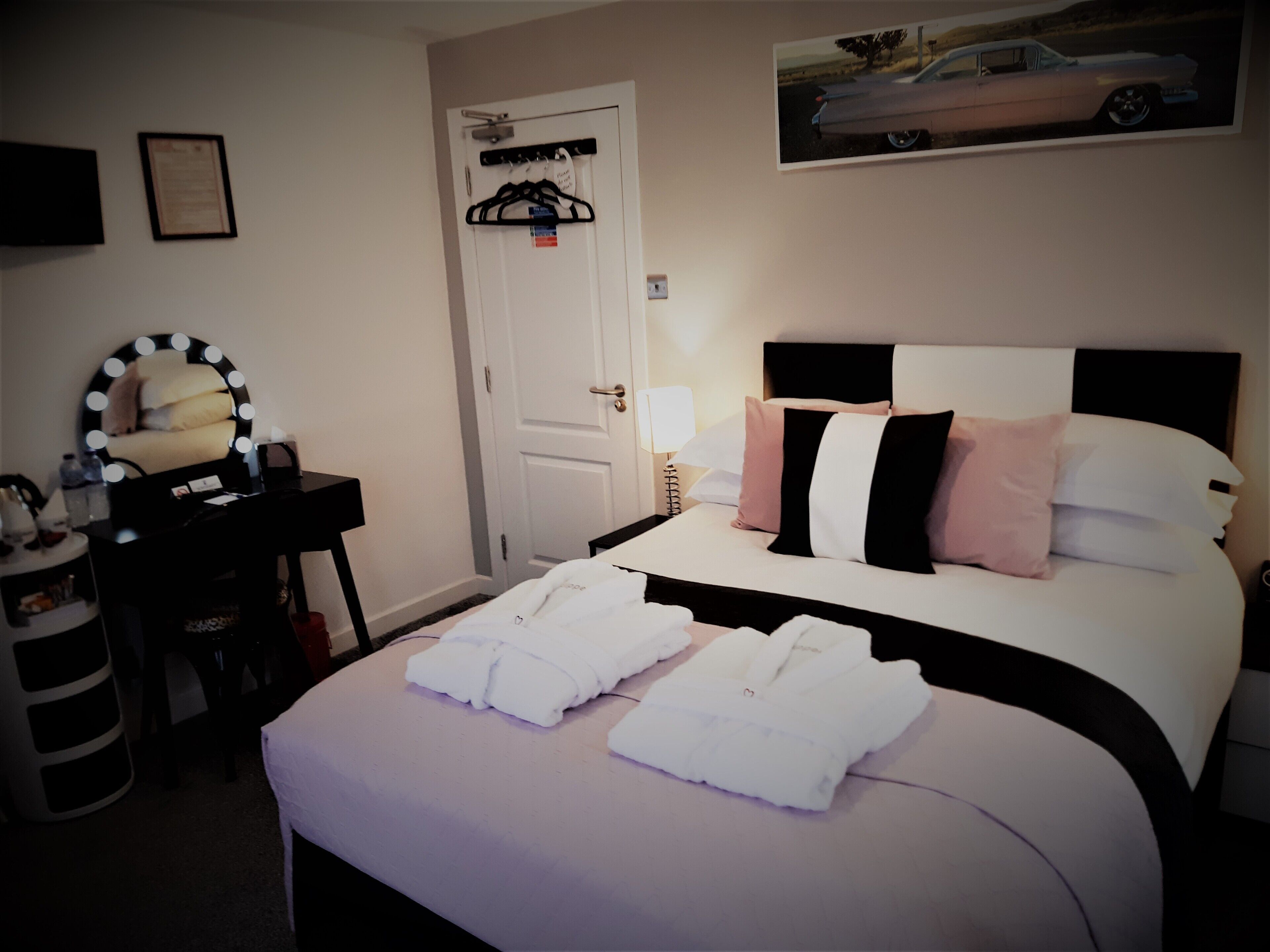 A hotel room with a bed, a vanity with a mirror, a coat rack, and a framed picture on the wall.