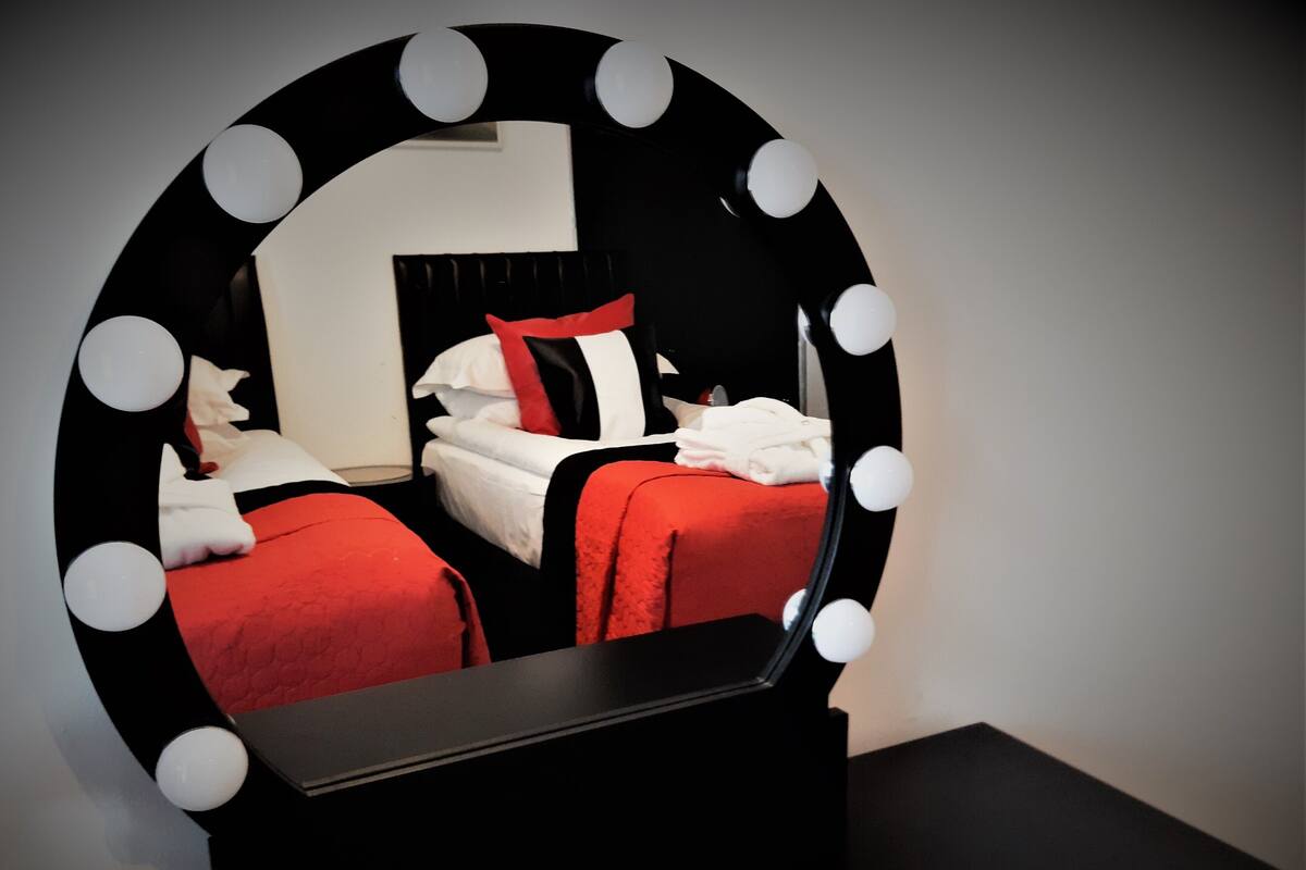 A room with a large mirror surrounded by lights, two beds with red bedding, and a black dresser.