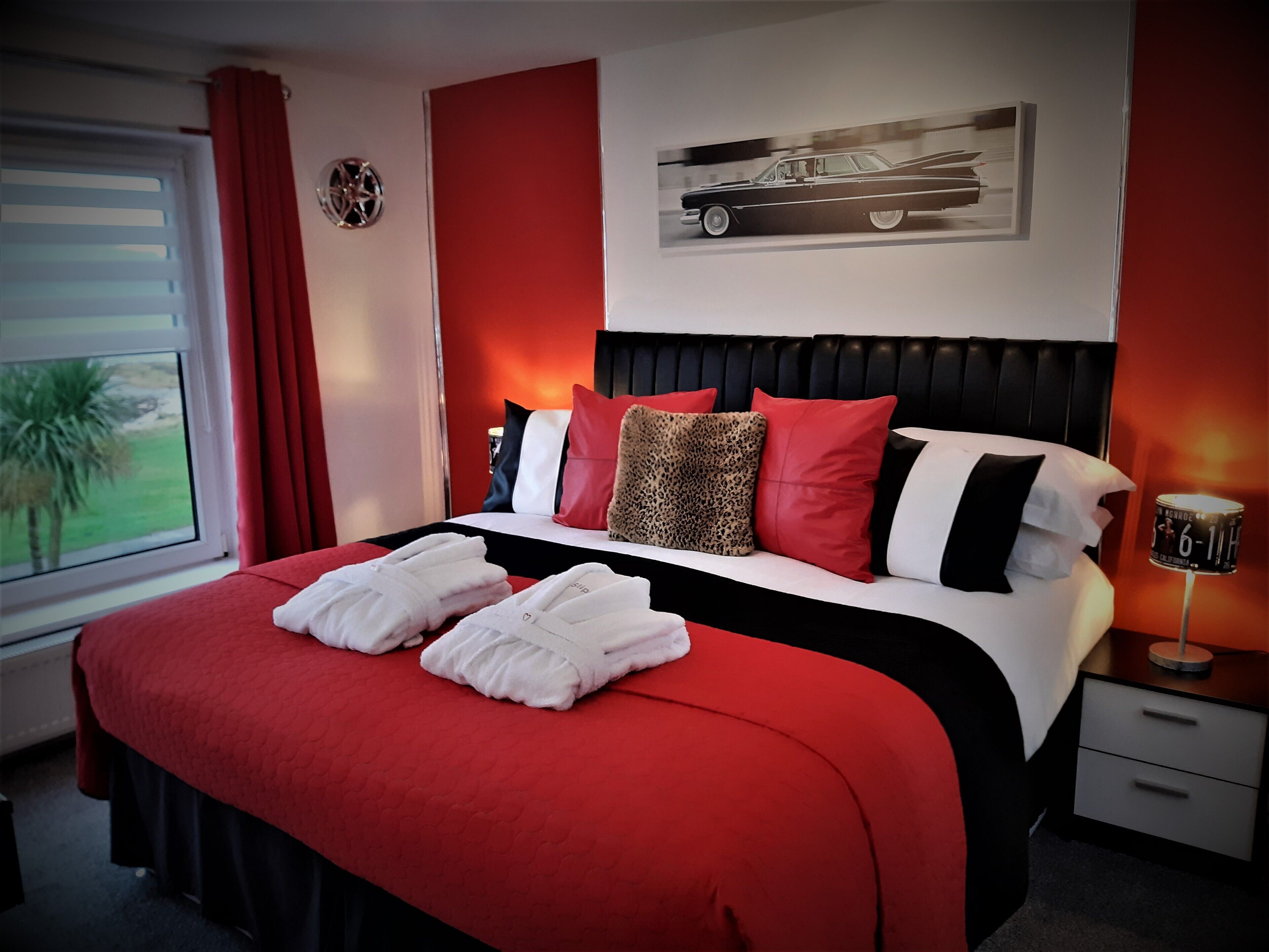 A hotel room with a bed, a nightstand, a lamp, a window with curtains, and a wall-mounted artwork of a car.