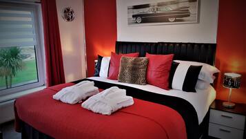 A hotel room with a bed, a nightstand, a lamp, a window with curtains, and a wall-mounted artwork of a car.