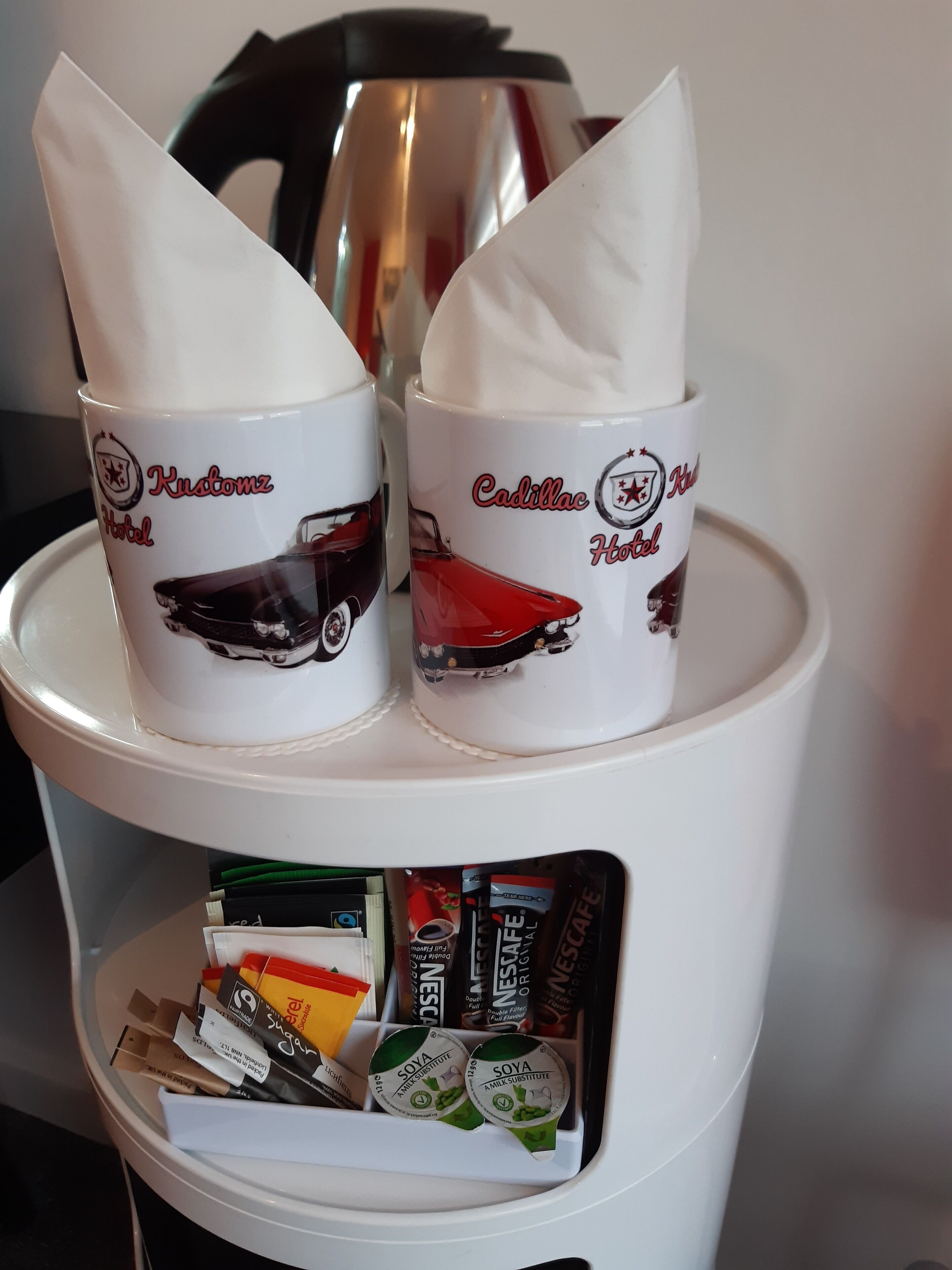 Dos tazas blancas con diseños de autos y nombres de hoteles sobre una estantería. Abajo, hay varias cápsulas de café y sobres.