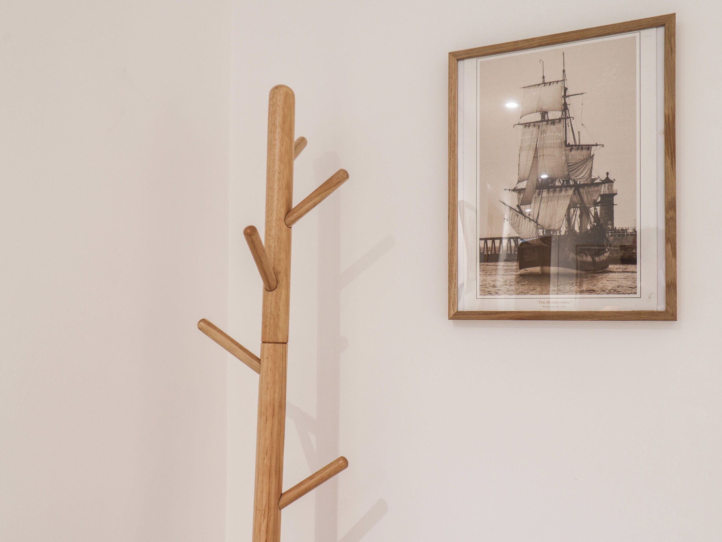 Un porte-manteau en bois, doté de crochets, est adossé à un mur blanc, à côté d’une photographie en noir et blanc encadrée représentant un navire.