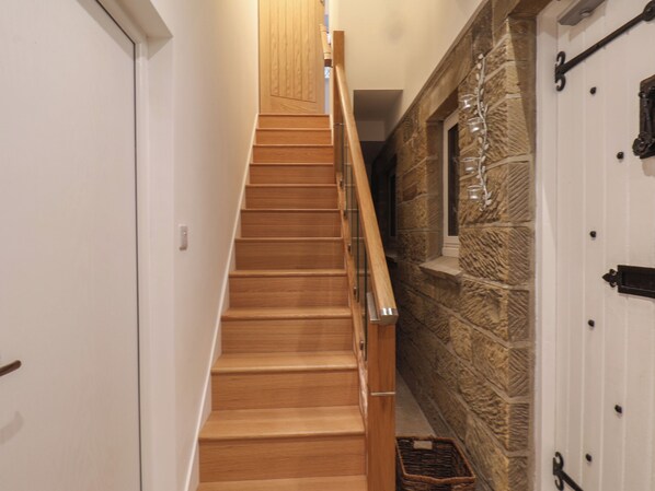 A wooden staircase with a handrail, leading to a room with a stone wall and a door with a number plate. - Brambles Apartment (Whitby)
