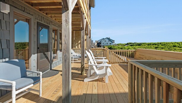 A wooden deck with white chairs and a sofa, a wooden railing, and a view of a house in the background. - Endless Summer: Direct Beach Access | Ocean Views (Duck)