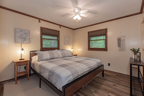 A bedroom with a bed, wooden floor, a ceiling fan, and two windows with blinds.