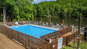 Una piscina rectangular con una cubierta de madera, rodeada por una valla de madera, y un lago al fondo.