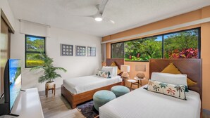 A hotel room with two beds, a TV, a ceiling fan, and a large window with a view of trees.