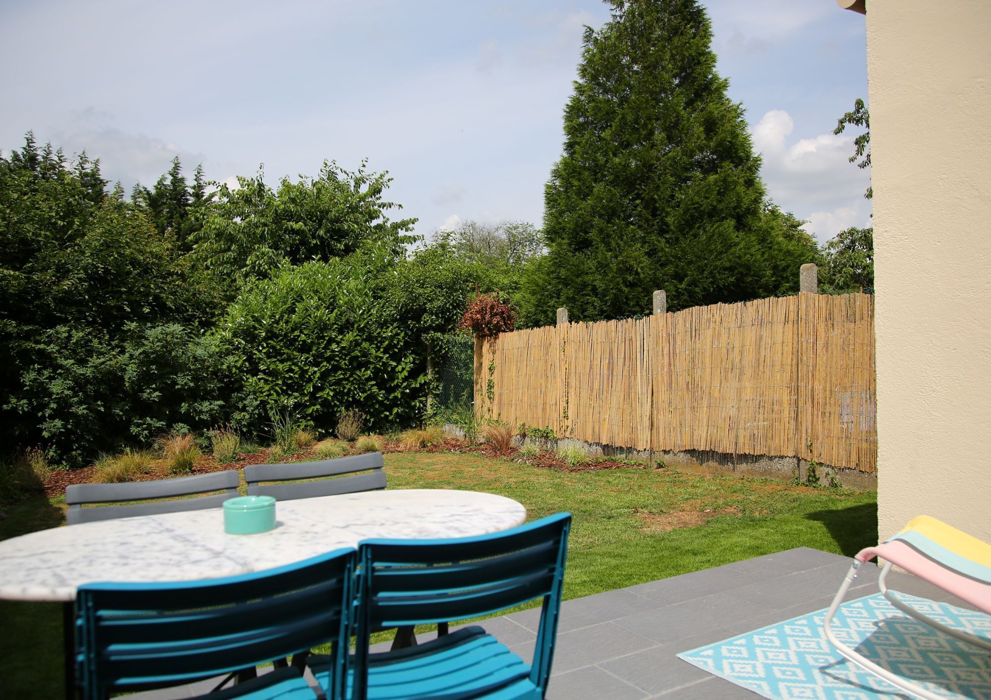 A patio with a round table and blue chairs, surrounded by a bamboo fence and lush greenery.