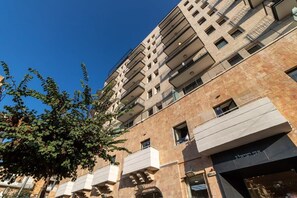 Um edifício com várias andares, varandas, céu claro e uma árvore em primeiro plano.