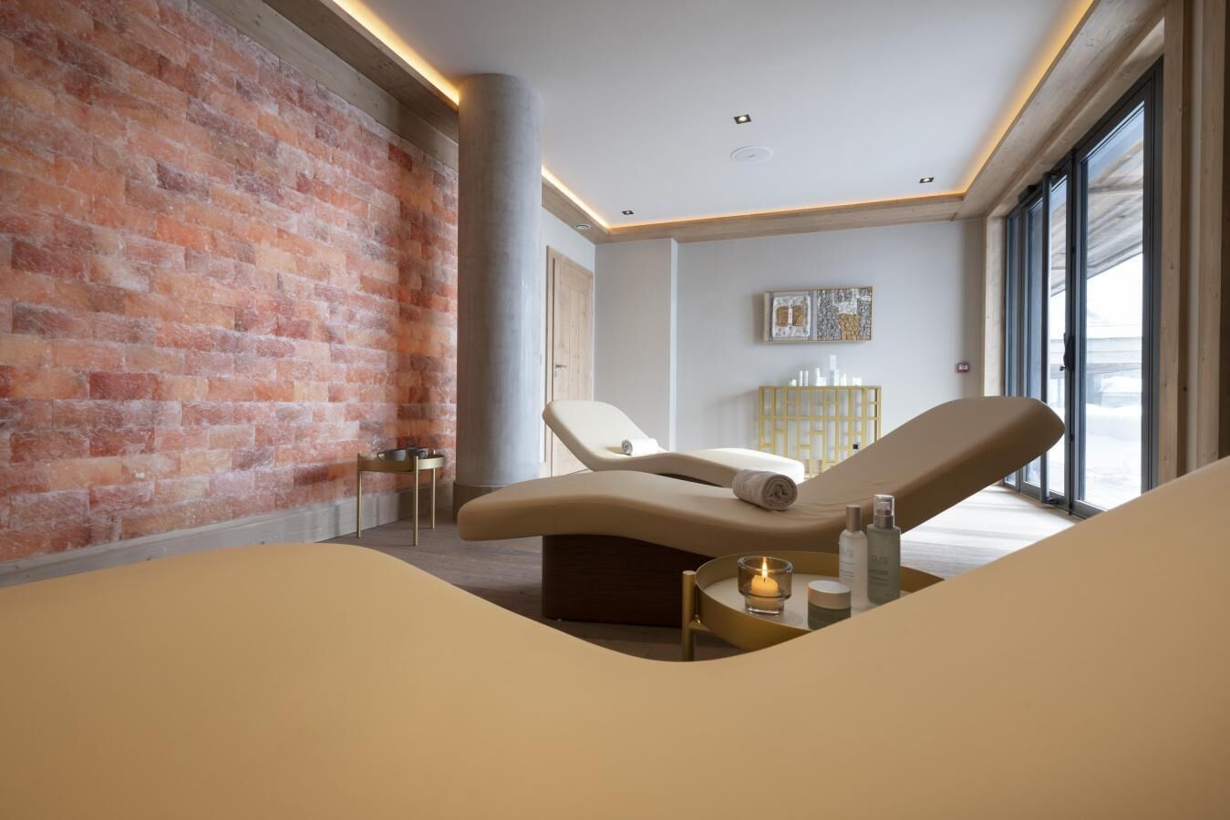 Une salle de spa avec un mur en briques, une table de massage, une petite table avec des bougies et une fenêtre donnant sur un paysage agréable.