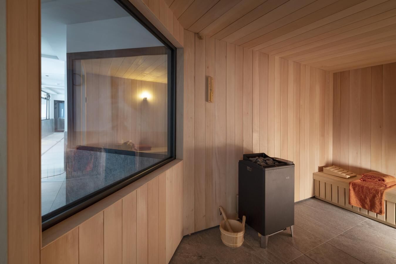 A sauna room with wooden walls, a large window, a sauna heater, and a wooden bench.