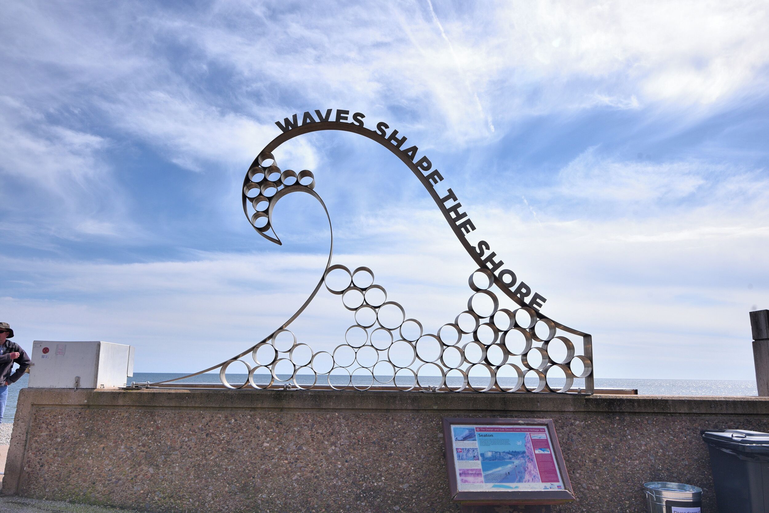Una scultura in metallo con il testo "WAVES SHAPE THE SHORE" sullo sfondo di un cielo sereno.