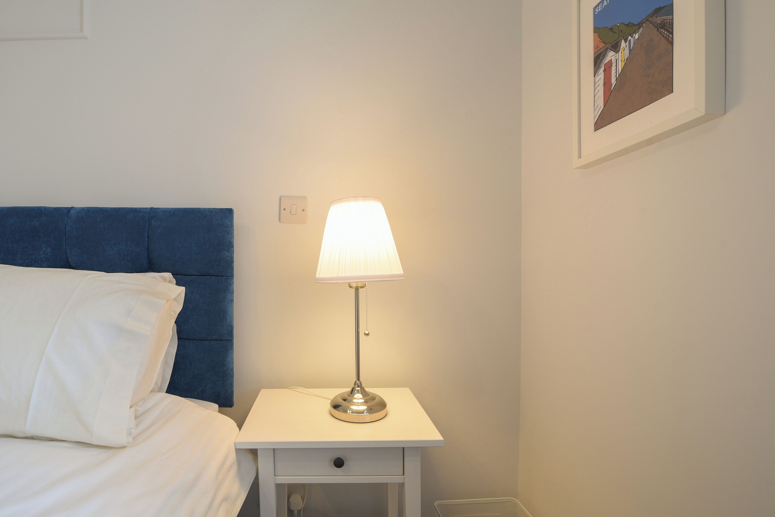 A neatly made bed with white linens, a blue headboard, a bedside table with a lamp, and a framed picture on the wall.