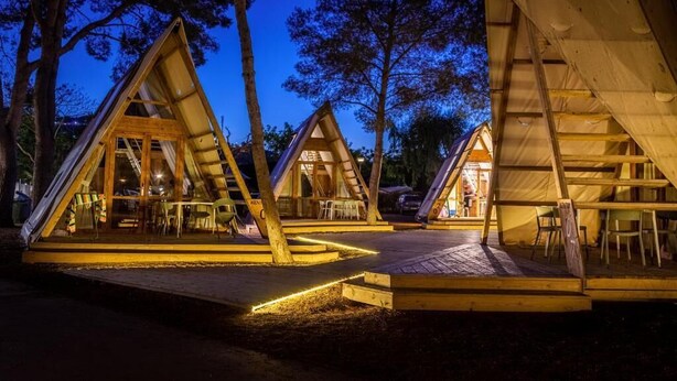 Three wooden triangular tents with interior seating and tables, set in a forested area at dusk. - Tamarit Beach Resort (Tarragona)