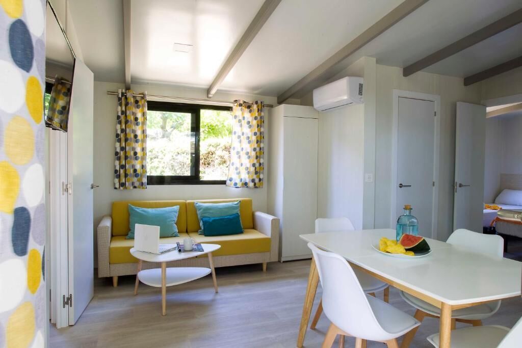 Una habitación luminosa con un sofá amarillo, una mesa blanca y un ventanal con cortinas.