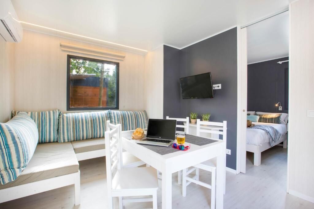 A modern, compact room with a white desk, a laptop, and a glass of beer. There is a bed with a striped pillow and a TV mounted on the wall.