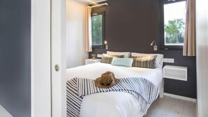 A modern bedroom with a large bed, striped bedding, a hat on the bed, and a window with curtains. - Tamarit Beach Resort (Tarragona)