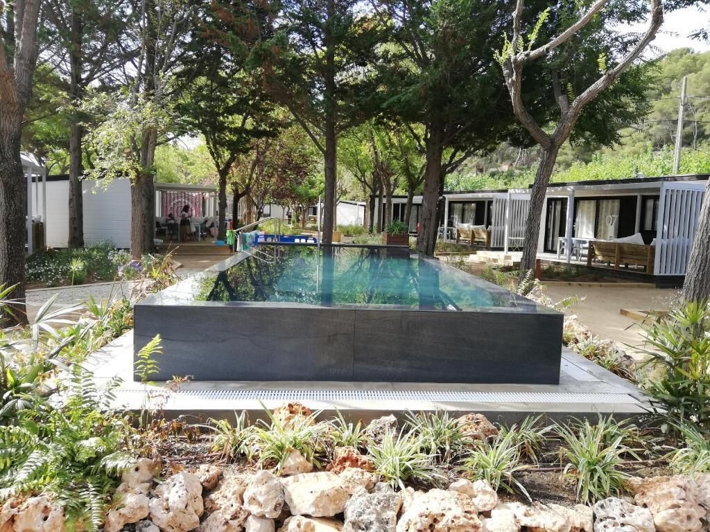 Een modern buitenzwembad met een rechthoekig zwembad, omgeven door rotsen en planten. Er staan houten ligstoelen en in de achtergrond is een overdekte zithoek.