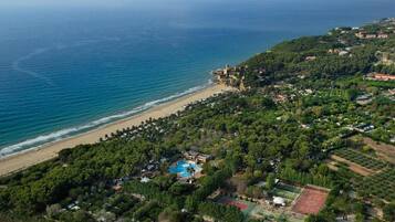 Vista aérea de una zona costera con una playa, una estructura que parece un castillo y una área residencial con piscina y canchas de tenis.