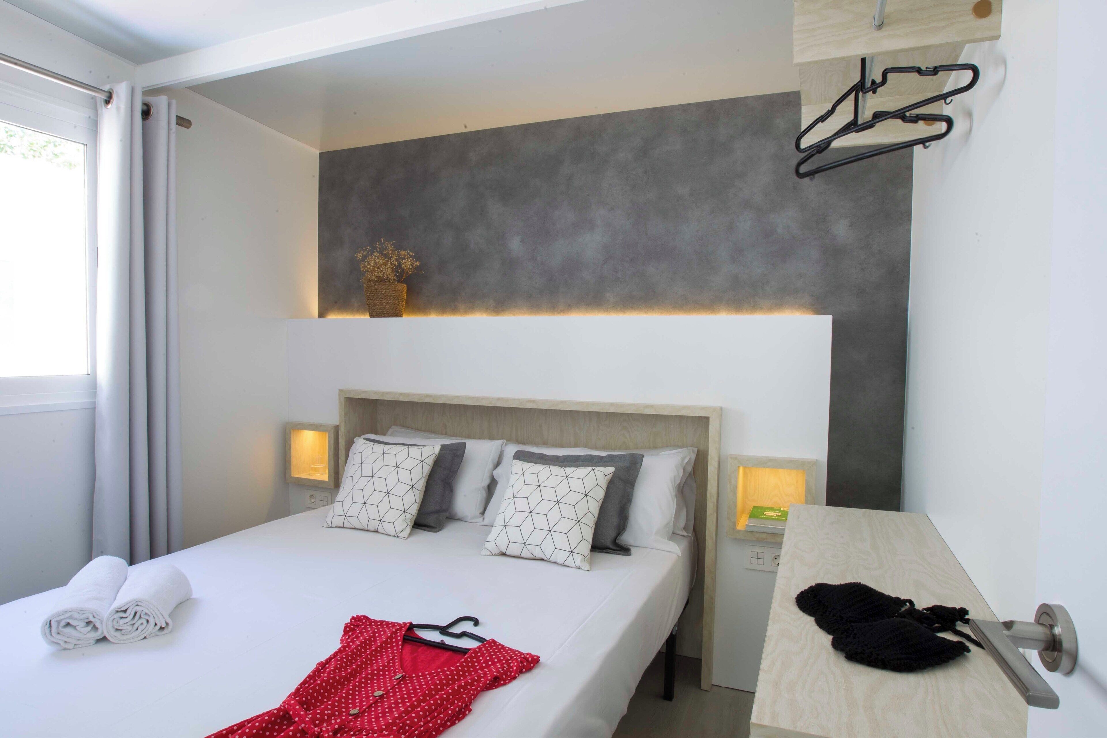 Una cama bien hecha con almohadas, una prenda roja y una prenda negra sobre una mesita de noche de madera.