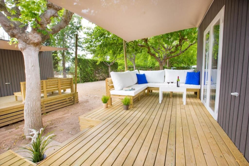 A wooden deck with a white sofa and blue cushions, surrounded by trees and a covered area.