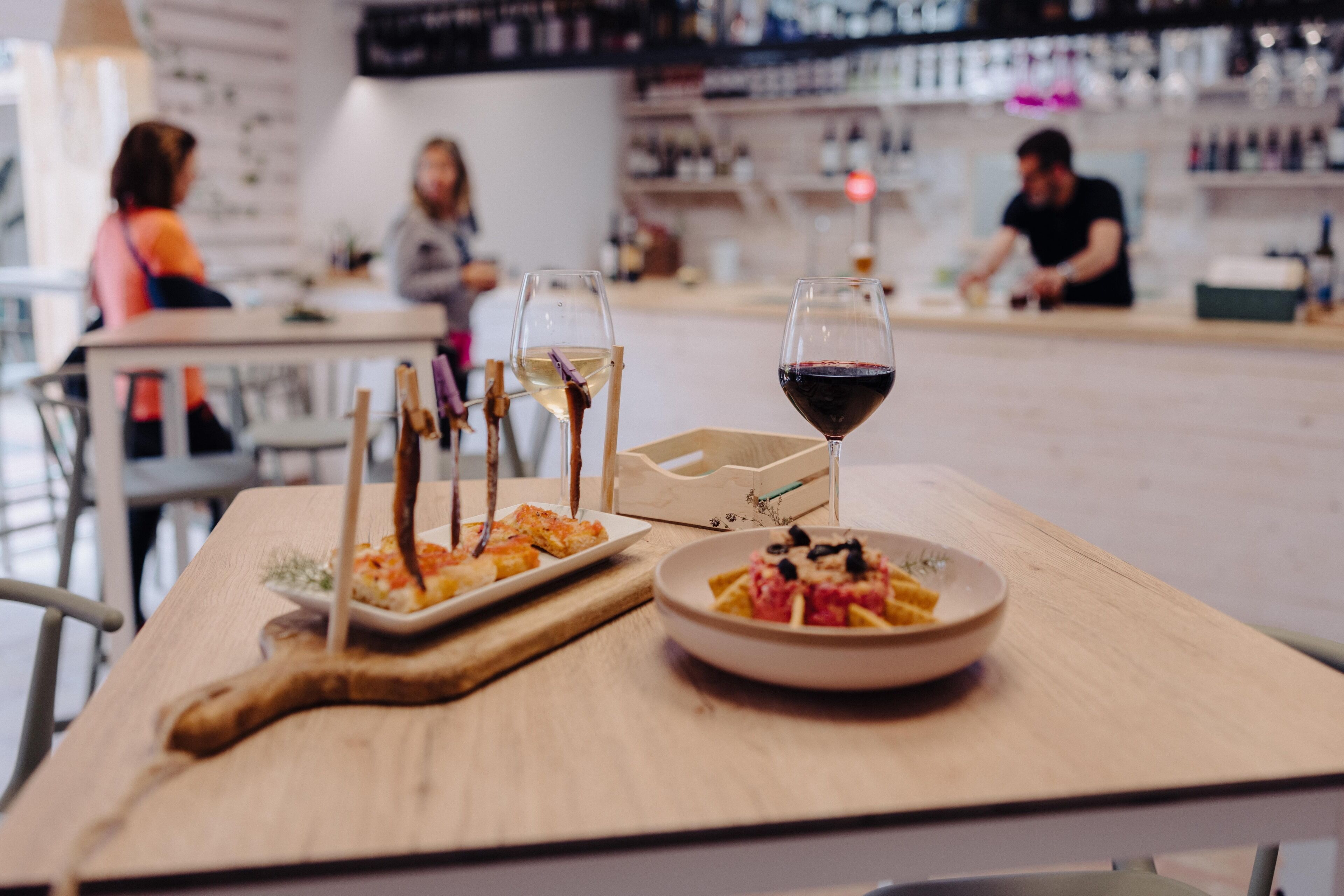 Un ambiente de restaurante con una mesa dispuesta para dos, que incluye una mesa de madera, dos copas de vino, un plato de pasta y una tabla de madera para servir.