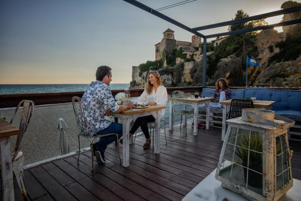 Una terraza de madera con mesas y sillas, con vistas a una zona costera y un edificio histórico.