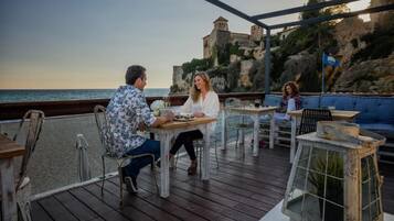 Una terraza de madera con mesas y sillas, con vistas a una zona costera y un edificio histórico.