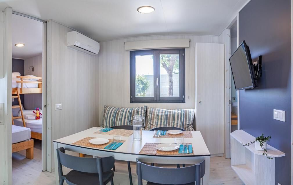 Habitación compacta con una mesa de comedor preparada para dos personas, una cama litera y un televisor montado en la pared.