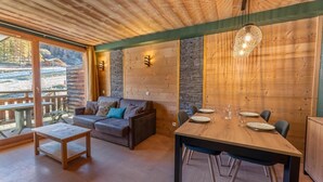 L’intérieur chaleureux d’un chalet avec une table à manger en bois, dressée pour quatre personnes, un canapé agrémenté de coussins et une grande fenêtre donnant sur une zone boisée.