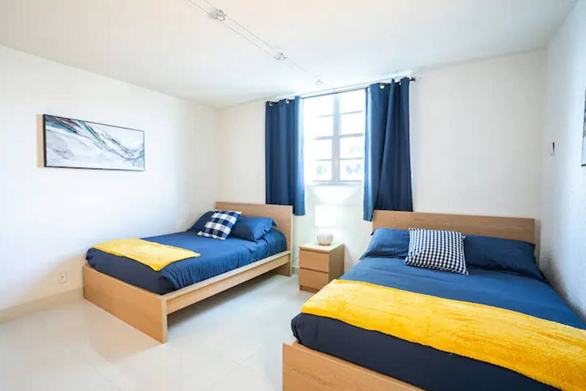A bedroom with two beds, a nightstand, a window with curtains, and a painting on the wall.