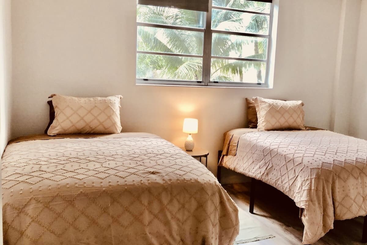Two beds with matching quilts, a nightstand with a lamp, and a window with a view of palm trees.