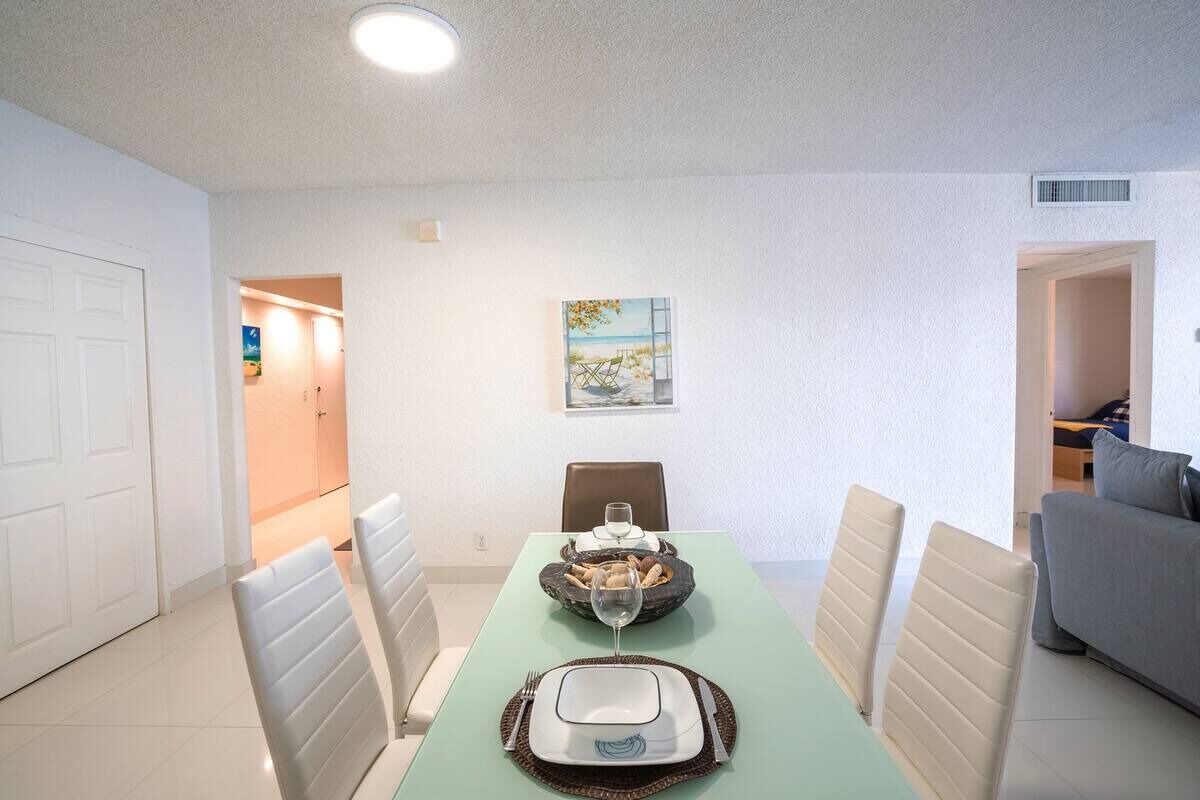 Zona de dinar amb taula preparada per a dues persones, cadires, un quadre a la paret i una porta que dona a una altra habitació.