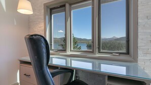 Un espacio de oficina moderno con un escritorio de vidrio, una silla de cuero y vistas a un lago y montañas a través de amplios ventanales.