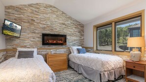 Un dormitorio con dos camas, una pared de piedra, una chimenea, un televisor y una ventana con vistas a árboles.