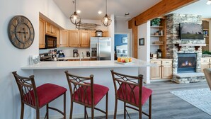A modern kitchen with wooden cabinets, a large island with bar stools, a stone fireplace, and a mounted TV. - Fourth & Main Condo: Heart of Frisco (Frisco)