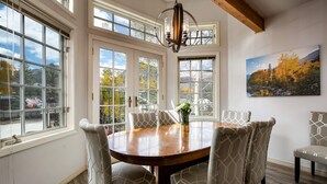 A dining area with a round wooden table, six patterned chairs, a chandelier, and large windows with a view of trees and mountains. - Fourth & Main Condo: Heart of Frisco (Frisco)