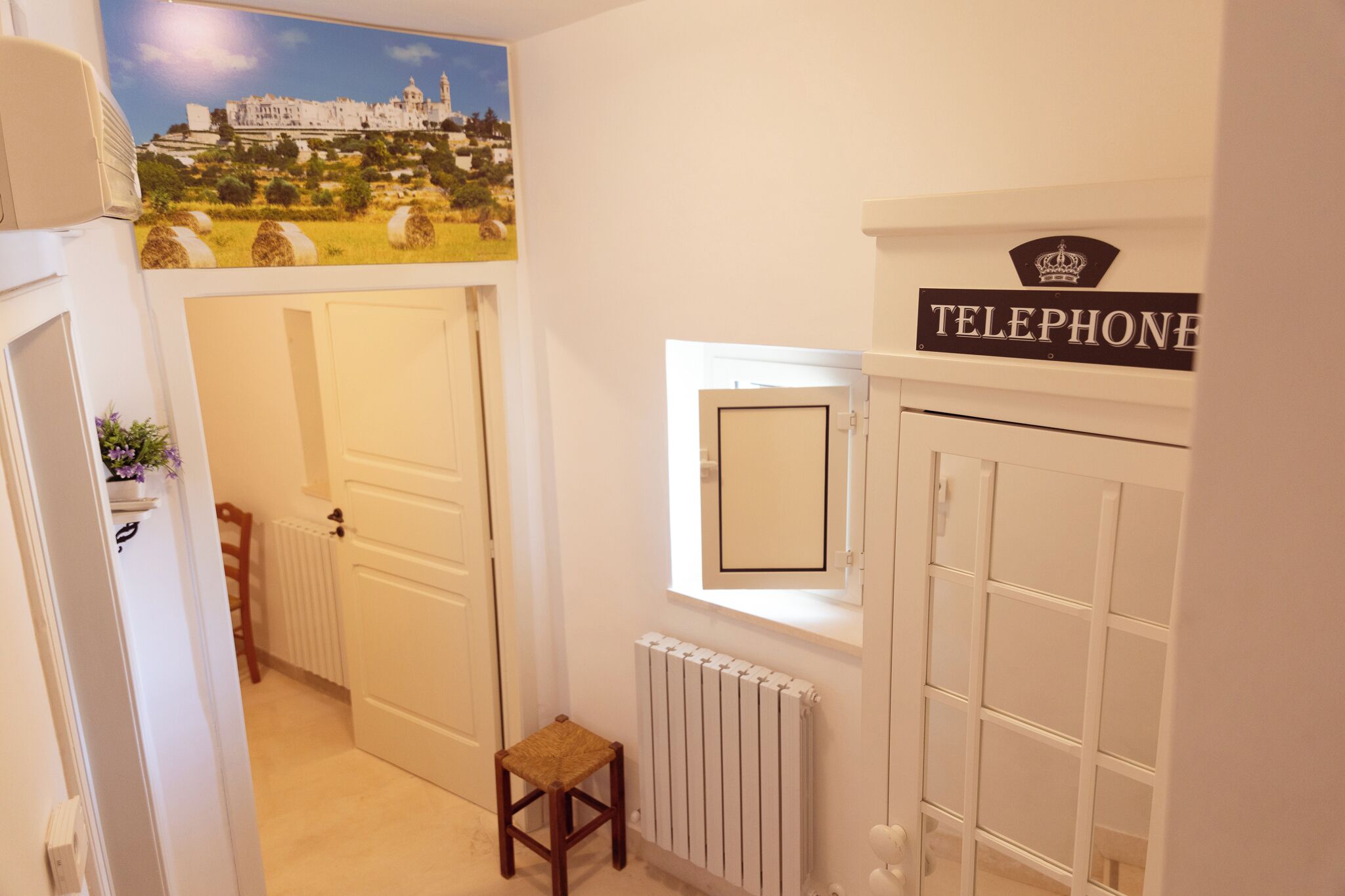 Un couloir avec un radiateur, un petit tabouret, une porte sur laquelle est apposée une pancarte indiquant « Téléphone », et un tableau encadré accroché au mur.
