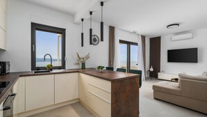 A modern kitchen with a central island, a dining area with green chairs, a beige sofa, and a large window offering a view of the sea. - 2-bedroom Villa La Vita with private heated 33sqm pool (Jesenice)