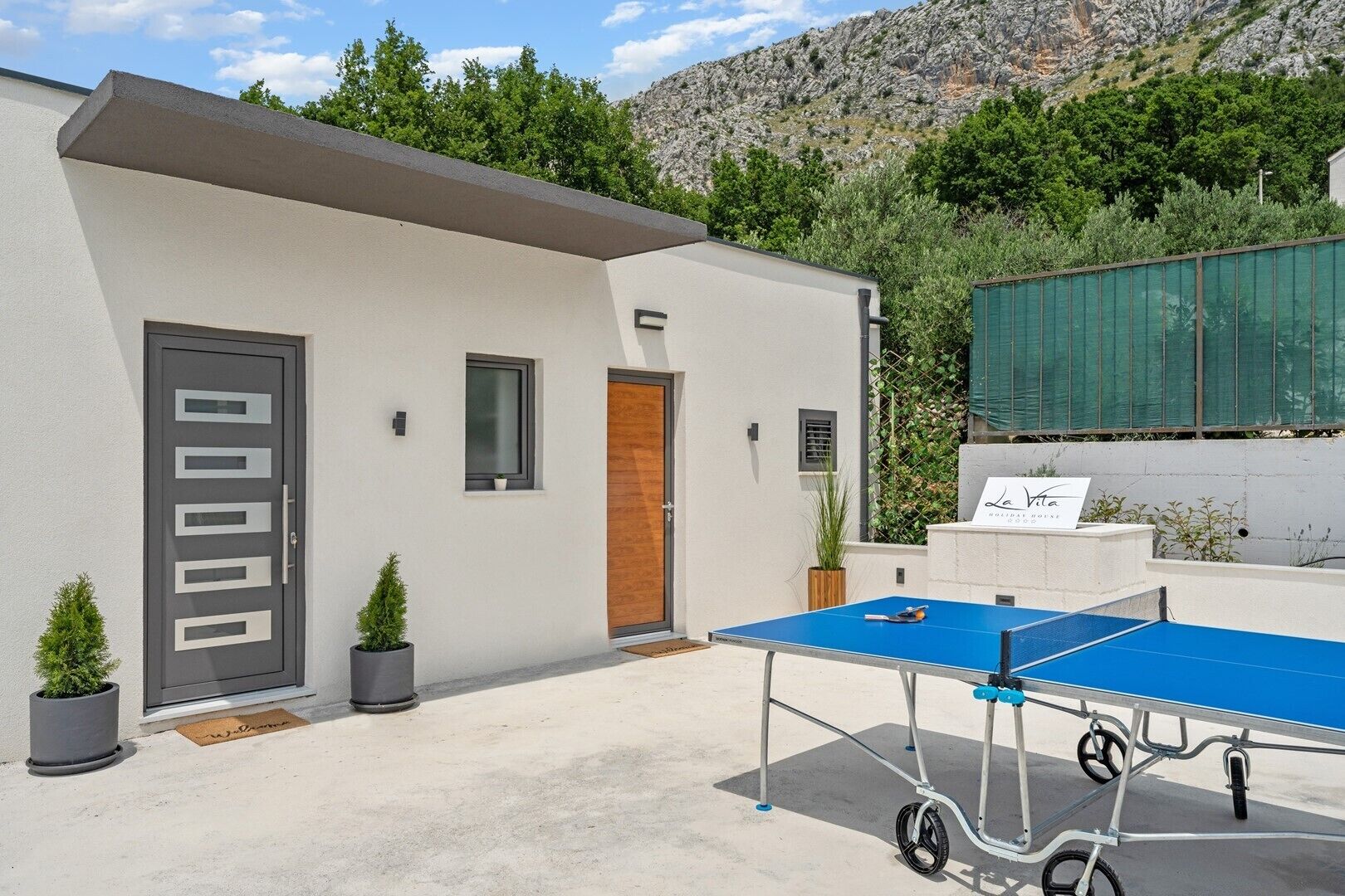 A modern building with a white exterior, a grey door with rectangular cutouts, a small window, and a blue ping pong table in the foreground.
