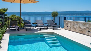 Zona junto a la piscina con sillones de descanso y una sombrilla, con vistas panorámicas al mar y las colinas.