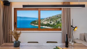 Un comedor con una mesa de madera, un jarrón con plantas secas y vistas a un paisaje costero a través de un ventanal grande.