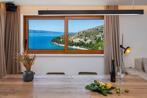 A dining area with a wooden table, a vase with dried plants, and a view of a coastal landscape through a large window.