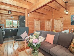 A cozy cabin interior with a wooden ceiling, a grey sofa with cushions, a wooden dining table, and a kitchen area with a sink and cabinets. - Pigsfoot Lodge (Tiverton)