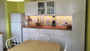 Een kleine keuken met witte kastjes, een koelkast, een tafel met een tafelkleed met een patroon en een hanglamp.