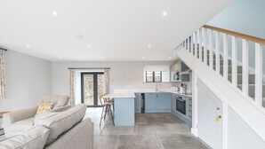 A modern kitchen with a central island, a dining area with a sofa and chairs, and a staircase with a wooden railing. - No 1 The Coach House (Carnforth)