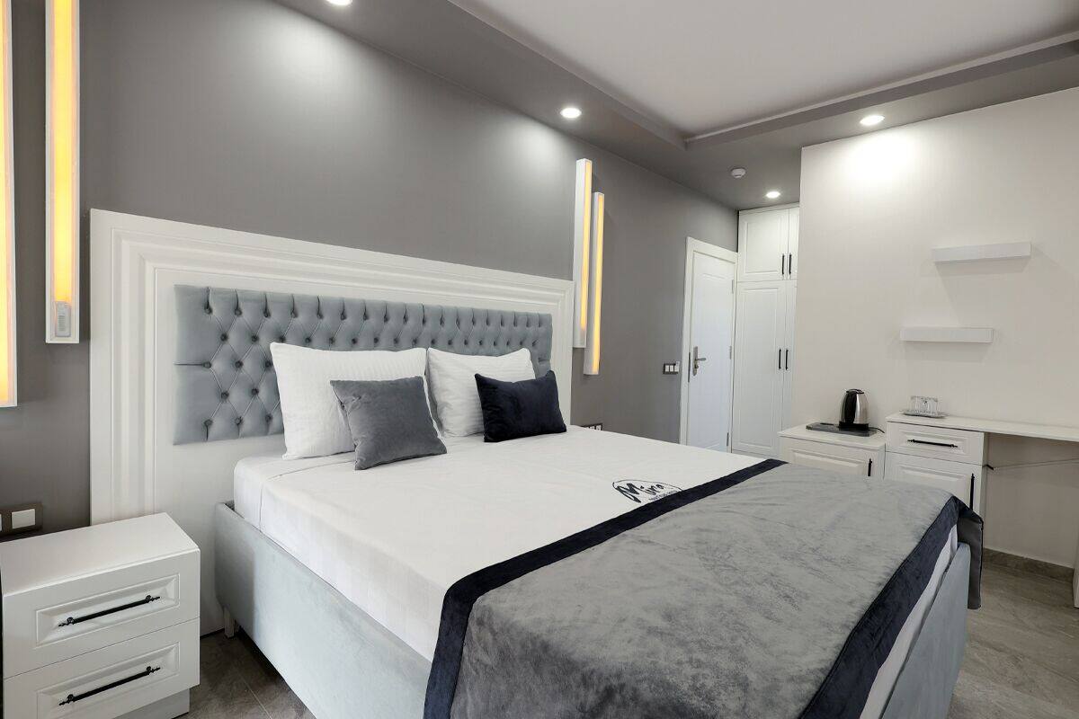 Una camera da letto moderna con una testiera trapuntata, un letto grande con biancheria da letto bianca e una coperta grigia, un comodino con una lampada e un armadio.