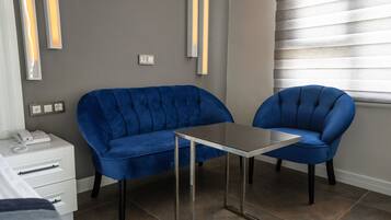Een moderne kamer met een blauwe bank en een bijpassende fauteuil, een klein tafeltje, een nachtkastje met een telefoon, en een raam met zonwering.